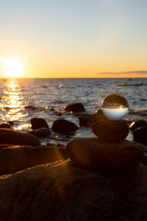 Glass ball lies on the top stone of stacked stones, at sunset on the beach, the sea and the setting sun are reflected in the ballの写真素材