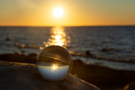 Glass ball on a rock at sunset on the beach, the sea and the setting sun are reflected in the ballの写真素材