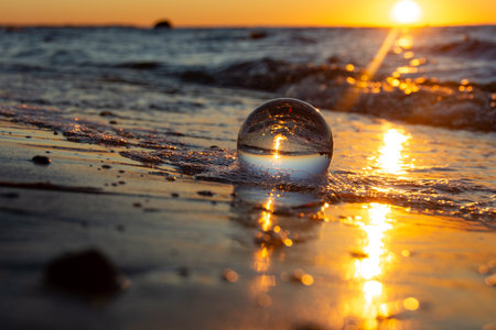 A glass ball lies in the waves on the sandy beach, the sea and the setting sun are reflected in the ballの写真素材