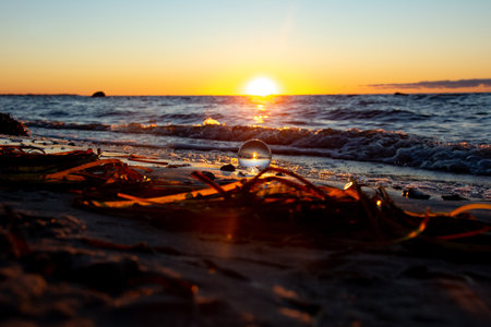 A glass ball lies in the sand in front of the waves, beach with algae and the sea and the setting sun are reflected in the ballの写真素材