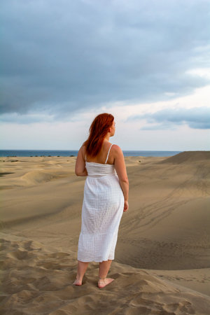 Young woman with red hair stands on a sand dune and looks at the seaの写真素材
