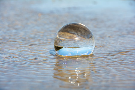 A glass ball lies in the waves on the sandy beach, the sea and the setting sun are reflected in the ballの写真素材