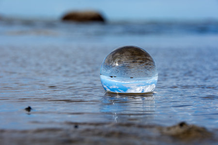 A glass ball lies in the waves on the sandy beach, the sea and the setting sun are reflected in the ballの写真素材