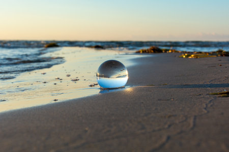 A glass ball lies in the waves on the sandy beach, the sea and the setting sun are reflected in the ballの写真素材