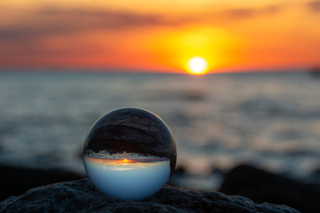Glass ball on a rock at sunset on the beach, the sea and the setting sun are reflected in the ballの写真素材