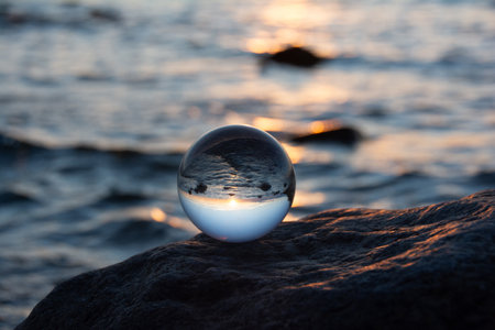 Glass ball on a rock at sunset on the beach, the sea and the setting sun are reflected in the ballの写真素材