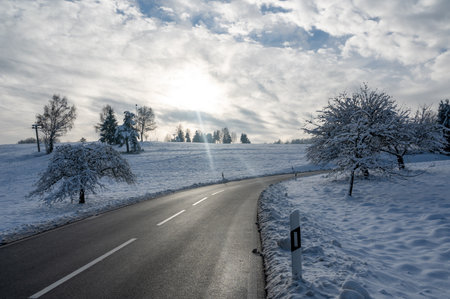 Snow landscape with trees, road and blue sky with clouds and sunの写真素材