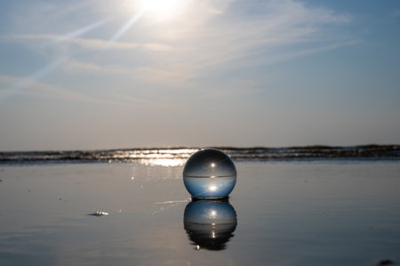 A glass ball lies in the waves on the sandy beach, the sea and the setting sun are reflected in the...