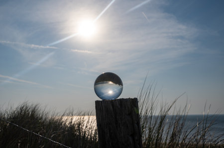 Glass ball lies on a wooden post, beach grass in the background, the sea, the sun and the blue sky are reflected in the ballの写真素材