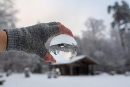 Human hand with a grey glove holds a glass ball reflecting a snow-covered landscape and a hutの写真素材