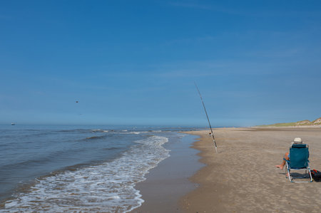A fisherman from behind, sitting on a folding chair on the sandy beach with his fishing rod by the seaの写真素材