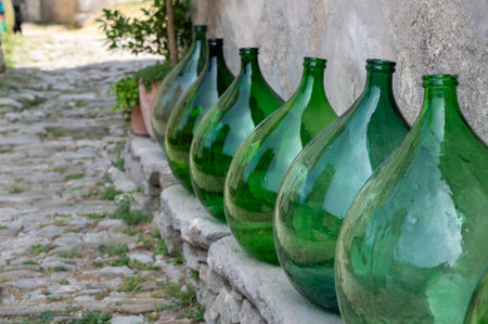 Some large green bulbous bottles stand on a low old wall on a house wall with part of an alley