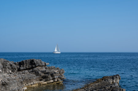 A sailing ship in the blue sea, rocks in the foreground and with blue skyの写真素材