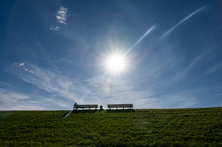 A person sits alone on a park bench on the horizon of a large green meadow, with bright sun and blue skyの写真素材