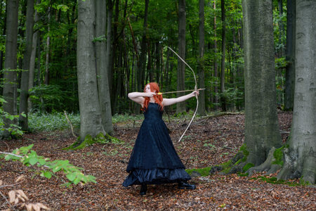 A young, red-haired woman, wearing a long black dress, is shooting with a bow and arrow, looking...