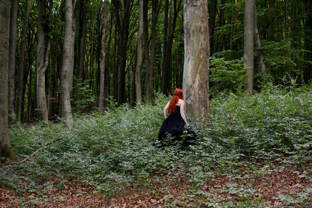 A red-haired woman in a long black dress stands in a green forest among ferns.