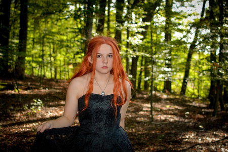 Beautiful young woman with long red hair and black dress is standing in the green forest, holding...