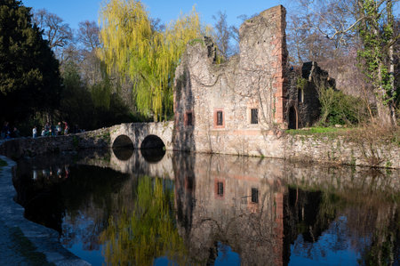 Old ruins by a small lake in a park, with an arched bridge and reflections in the water, in...