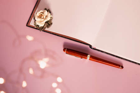 A flat lay of a blank notebook with a rose on it, a wooden pen and some blurred fairy light on a pink backgroundの写真素材