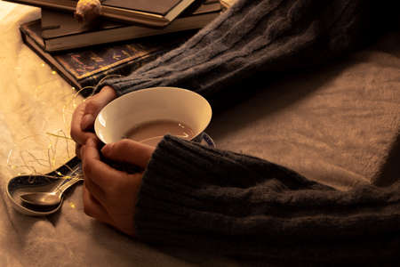 An unrecognizable girl is holding a hot cup of tea, placed next to a teaspoon. There are some books on the backgroundの写真素材