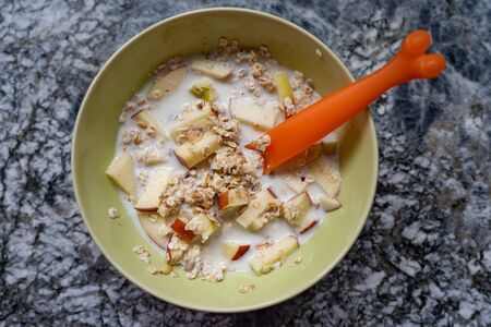 Preparing a Swiss Muesli with Fresh Milk, Cereals, Dry Grapes and Apples in a Light Green Bowl and Orange Spoon on a Granite Table in the Kitchen by Daylight. High angle, closeup.の写真素材