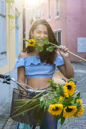 Beautiful young lady biting in sunflower while walking through the old city and pushing her turquoise bicycle. Sunbeam at background.の写真素材