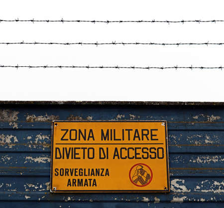 Military area with yellow sign in Italian language: "Military zone, no entry, armed surveillance". Translation: "Military zone, no entry, armed surveillance".の写真素材