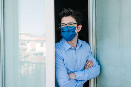 Young businessman with mask forced to stay in quarantine at home for Corona virus looking out the window.の写真素材