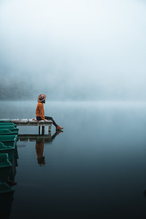 Side view of fashioned young woman sitting on wooden dock looking at view on a misty morning. Female...