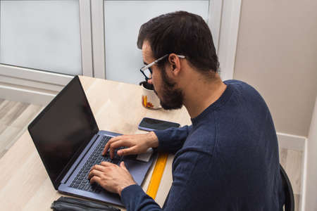 Young caucasian man in a blue sweater and glasses working from home while talking to a colleague and having a cup of coffee / tea with a laptop and a tableの写真素材