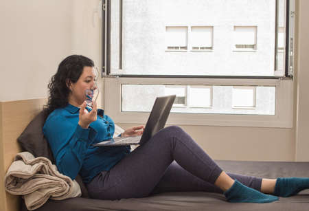 Young caucasian woman working from home on her bed in a blue formal shirt and night pants and...