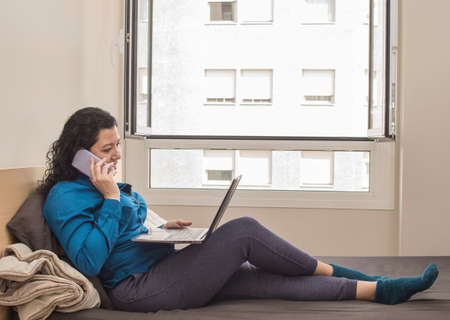 Young caucasian woman working from home on her bed in a blue formal shirt and night pants and stockings while talking on her mobile work phone and on the right a windowの写真素材