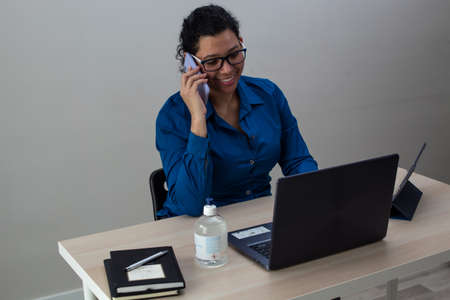 Young caucasian woman in a blue shirt and glasses working from home on her laptop while talking on the phone with a friend on a table with black books and a hand sanitizerの写真素材