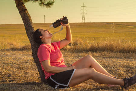 Young athletic woman resting on a tree after running wearing sports clothing, striped shirt and...
