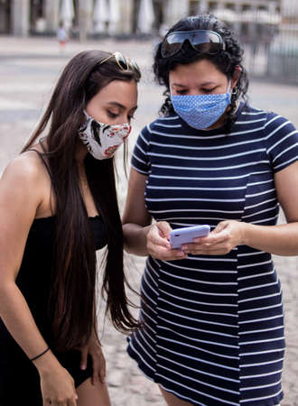 Selective focus of young women walking around a square in the city using medical masks while using their electronic devicesの写真素材