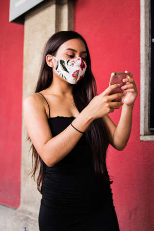 Selective focus of young Hispanic woman walking around a square while using her smartphone and wearing her medical maskの写真素材