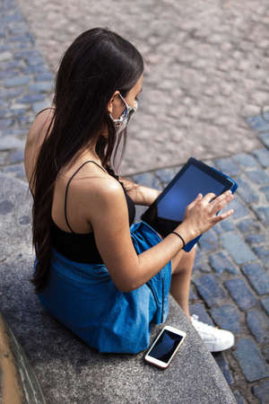Selective focus of young hispanic woman strolling around a square in the city while using her electronic devices, her smartphone and tabletの写真素材
