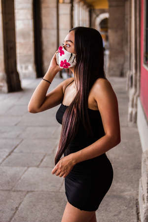 Selective focus of young Hispanic woman walking around a square while using her smartphone and wearing her medical maskの写真素材