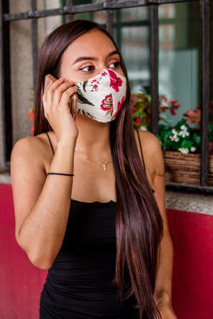 Selective focus of young Hispanic woman walking around a square while using her smartphone and wearing her medical maskの写真素材