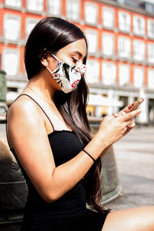 Selective focus of young Hispanic woman walking around a square while using her smartphone and wearing her medical maskの写真素材