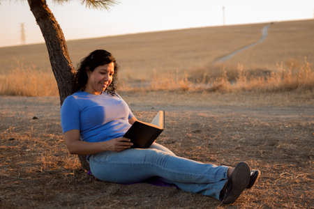 Young Caucasian woman reading a black book sitting on the ground in a park with sunset in the backgroundの写真素材