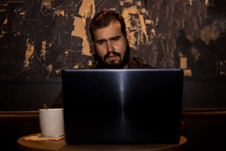 Young caucasian man with a black shirt in a shop working with his laptop having a coffee and with his phoneの写真素材