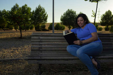 Caucasian woman sitting on a bench in the park reading a black book while wearing jeans and a blue blouseの写真素材