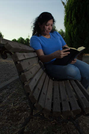 Caucasian woman sitting on a bench in the park reading a black book while wearing jeans and a blue blouseの写真素材