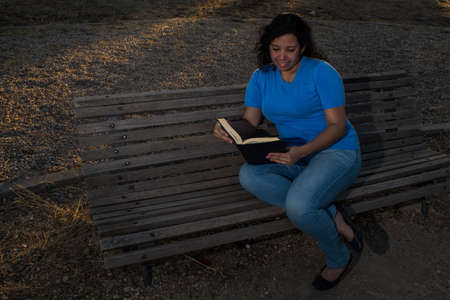Caucasian woman sitting on a bench in the park reading a black book while wearing jeans and a blue...