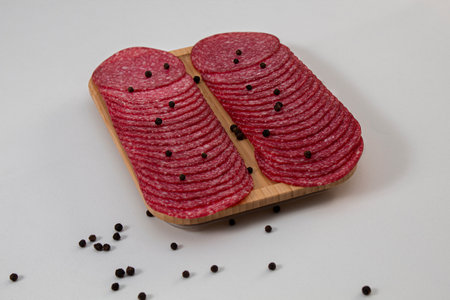 Slices of salami on a wooden board underneath whole slices of black pepper with a white backgroundの写真素材