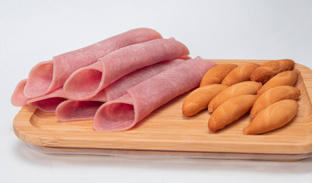 Slices of smoked ham with slices of bread on a wooden board with a white background and a very illuminated baseの写真素材