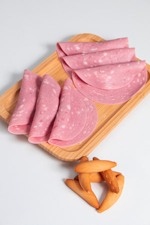 Slices of mortadella accompanied by slices of bread on a wooden board with a white background and a very bright baseの写真素材