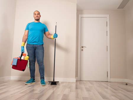 Young man cleaning with a sweeping brush and mop and bucket of water and cleaning supplies at home wearing blue gloves and blue shirtの写真素材