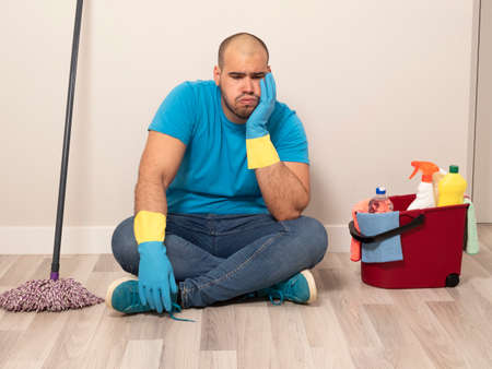 Young man cleaning with a sweeping brush and mop and bucket of water and cleaning supplies at home wearing blue gloves and blue shirtの写真素材